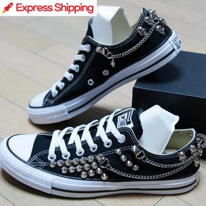 Puede incluir: Zapatillas bajas de lona negras con tachuelas y cadenas plateadas. Las zapatillas tienen cordones blancos y una suela de goma blanca.