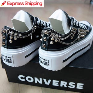 Puede incluir: Zapatillas Converse negras con plataforma y detalles de tachuelas y cadenas de metal plateado. Las zapatillas tienen suelas y cordones blancos, y el logo de Converse All Star es visible en el talón. Las zapatillas están sobre una caja negra de Converse.