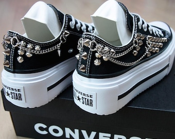 May include: Black Converse platform sneakers with silver metal studs and chain accents. The sneakers have white soles and laces, and the Converse All Star logo is visible on the heel. The shoes are on a black Converse box.