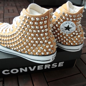 Puede incluir: Un par de zapatillas Converse de caña alta de color marrón claro con tachuelas piramidales plateadas que cubren toda la zapatilla. Las zapatillas están sobre una caja negra con el logotipo de Converse.