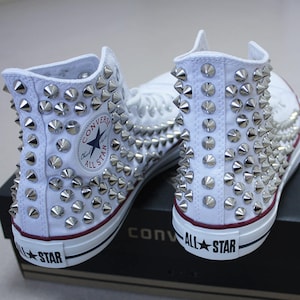 Puede incluir: Zapatillas Converse blancas de caña alta completamente cubiertas de tachuelas piramidales plateadas. Las zapatillas tienen una franja roja alrededor del borde de la suela y las palabras "All Star" en negro en la parte posterior de la zapatilla.