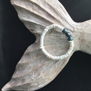 May include: A white shell bracelet with three dark gray pearls. The bracelet is made of small, flat, white shells strung together. The pearls are a dark gray color and add a touch of elegance to the bracelet. The bracelet is displayed on a wooden, fish-shaped carving.