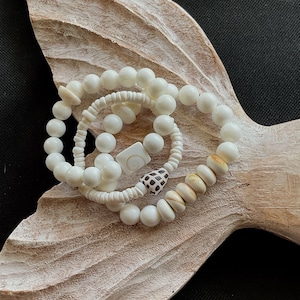 May include: Three white shell bead bracelets stacked on a white wooden mermaid tail. The bracelets are made of white and cream colored beads with a small brown and white shell accent.