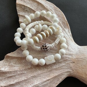 May include: Three white shell bracelets with different sized beads and shell accents. The bracelets are stacked on top of each other on a light brown wooden surface.