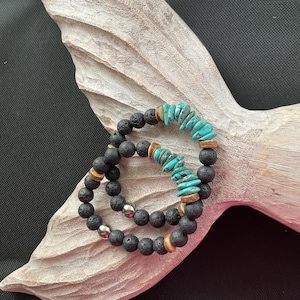 May include: Two bracelets made of black lava rock beads, turquoise stones, and wooden beads. The bracelets are stacked on top of each other.