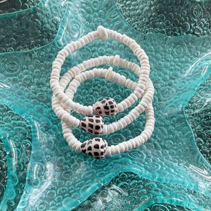 May include: Three white shell bracelets with black and white shell accents. The bracelets are stacked on top of each other and are lying on a blue and white textured surface.