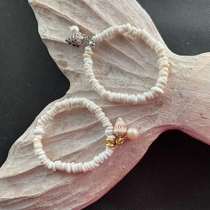 May include: Two bracelets made of small, off-white disc-shaped beads. One bracelet features a small, speckled seashell charm and a white pearl bead. The other bracelet includes a small, conical seashell charm and a round, pale pink pearl bead.