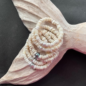 May include: A stack of five white shell rings with small, irregular shell pieces. Each ring is adorned with a few small, iridescent pearls. The rings are displayed on a light-colored, textured wooden surface.