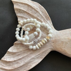 May include: Three white shell bead bracelets with a white and brown shell charm. The bracelets are on a white wooden surface.