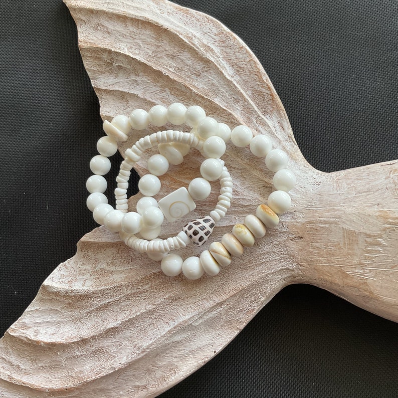 May include: Three white shell bead bracelets with a white and brown shell charm. The bracelets are on a white wooden surface.