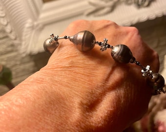 May include: A beaded bracelet featuring large, dark gray, round beads with a striped pattern. The beads are connected by smaller black beads and silver-toned accents. The bracelet is shown on a person's wrist.