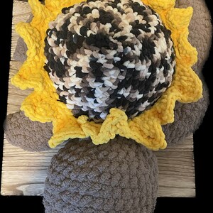 May include: A crocheted sunflower pillow with a brown center and yellow petals. The pillow is sitting on a wooden surface.