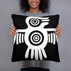 May include: Black throw pillow featuring a white graphic of a stylized bird. The bird design has concentric circles and lines, creating a bold, graphic look. The pillow is held by a person, and the background is a solid gray.