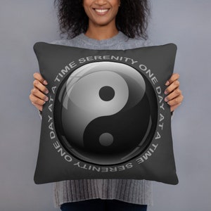 Könnte beinhalten: Ein schwarzes quadratisches Kissen mit einem Yin-und-Yang-Symbol in Grautönen. Das Design ist von dem Text "ONE DAY AT A TIME SERENITY" in hellgrauer Schrift umgeben. Das Kissen wird von einer Person gehalten.