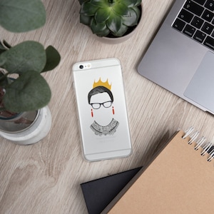May include: Clear phone case featuring a graphic of a woman with glasses, a crown, and a decorative collar. The design includes black, gold, and red accents. The phone case is on a wooden surface with a laptop, notebook, and plants.