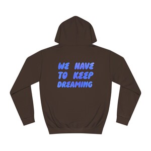 Può includere: Una felpa con cappuccio marrone con il testo "WE HAVE TO KEEP DREAMING" in blu sul retro.
