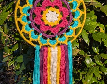 Puede incluir: Un atrapasueños hecho a mano con un aro amarillo y un diseño de ganchillo colorido. El ganchillo presenta detalles turquesa, amarillo, morado, rosa y blanco. Debajo, cuelgan hebras de hilo de colores, agregando textura e interés visual.