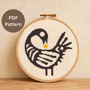 Könnte beinhalten: Kreuzstichmuster eines schwarz-weißen Vogels mit gelbem Detail, präsentiert in einem hölzernen Stickrahmen. Der Text "PDF Pattern" befindet sich in einem braunen Kreis. Das Design befindet sich auf beigem Stoff, ideal für Bastelprojekte.