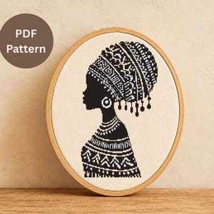May include: A black and white cross-stitch pattern of a woman in profile, wearing a detailed head wrap and jewelry, displayed in an oval wooden embroidery hoop. The text "PDF Pattern" is in a brown circle.