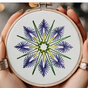 Könnte beinhalten: Gesticktes Blumendesign in einem Holzreifen. Das Kunstwerk zeigt ein symmetrisches Muster mit lila, grünen und gelben Fäden auf einem weißen Stoff. Der Reifen wird von Händen mit goldenen Ringen gehalten.