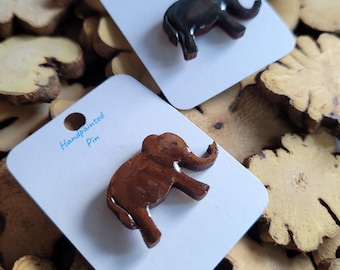 May include: Two hand-painted elephant pins on white cards. One pin is dark brown, the other is light brown. The cards are on a bed of wood slices. The text "Handpainted Pin" is visible on the cards.