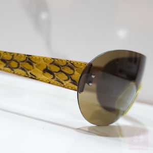 Peut inclure: Gros plan sur des lunettes de soleil avec une branche jaune &agrave; motif peau de serpent et un verre marron semi-circulaire. Les lunettes ont un design moderne, avec le verre fix&eacute; &agrave; la branche par deux petits rivets.