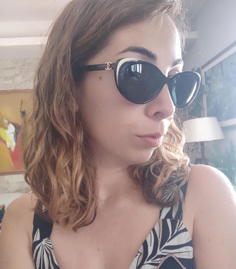 Puede incluir: Gafas de sol negras de gran tama&ntilde;o con un acento blanco en la parte superior de la montura. Las gafas de sol tienen lentes oscuras y un logotipo dorado en las patillas. La montura es de color marr&oacute;n oscuro. Las gafas de sol las lleva una persona.