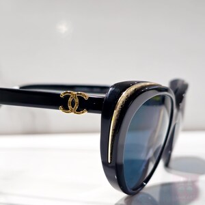 Puede incluir: Gafas de sol negras con un borde dorado y un logotipo dorado en la patilla. Las lentes son azules. Las gafas de sol tienen forma de ojo de gato. La montura es brillante.