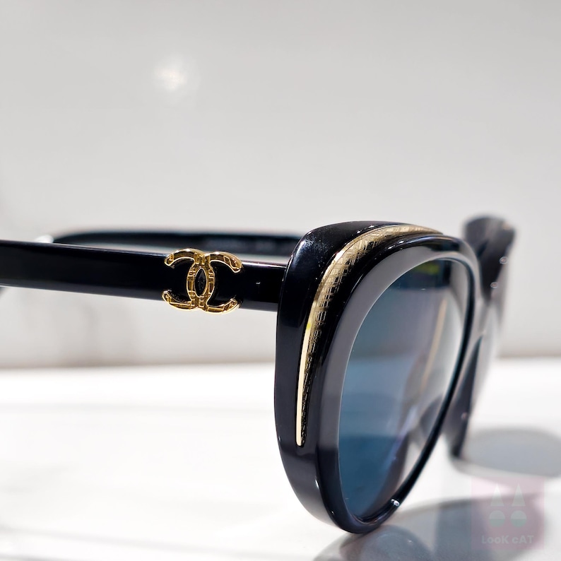 Puede incluir: Gafas de sol negras con un borde dorado y un logotipo dorado en la patilla. Las lentes son azules. Las gafas de sol tienen forma de ojo de gato. La montura es brillante.
