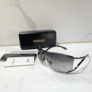 May include: Black Versace shield sunglasses with a gradient gray lens and a thin black frame. The set includes a black rectangular hard case with the gold Versace logo, a black cleaning cloth, and a white informational booklet with Versace text.