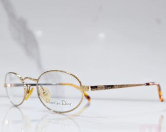 Puede incluir: Gafas ovaladas de color dorado con lentes transparentes. El nombre de la marca "Christian Dior" está impreso en la montura.