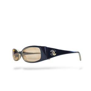 May include: Dark navy blue sunglasses featuring oval-shaped lenses with a light brown tint. The wide temples are adorned with a silver-toned interlocking CC logo emblem on the side, providing a distinct metallic detail against the dark frame.