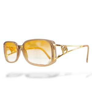 May include: Tan-colored sunglasses with a square frame and gradient yellow lenses. The temples are gold-colored with a decorative design. The sunglasses are set against a white background.