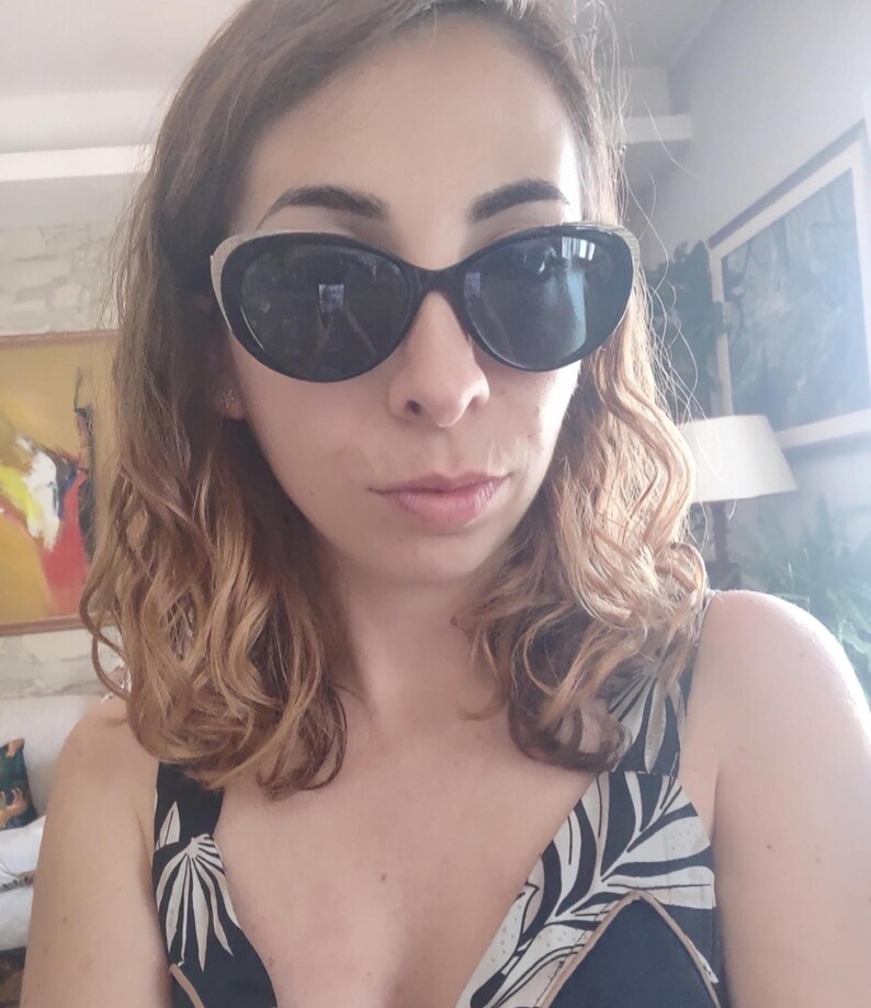 Puede incluir: Gafas de sol negras de ojo de gato con un acento plateado en la parte superior de la montura. La persona lleva una camiseta con estampado en blanco y negro. La persona tiene el pelo ondulado casta&ntilde;o claro.