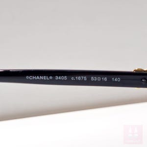 Puede incluir: Primer plano de la patilla de unas gafas Chanel negras. El texto en la patilla dice "CHANEL 3405 c.1675 53 16 140". Las gafas tienen detalles dorados.