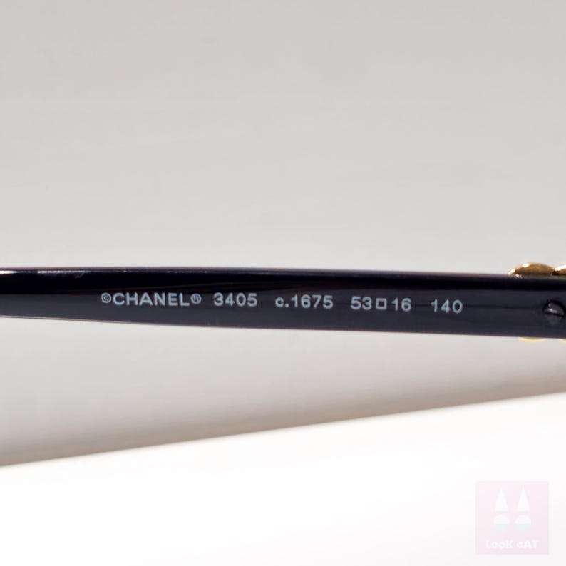 Puede incluir: Primer plano de la patilla de unas gafas Chanel negras. El texto en la patilla dice "CHANEL 3405 c.1675 53 16 140". Las gafas tienen detalles dorados.