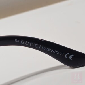 May include: Close-up of the arm of a black Gucci sunglass frame. The text "135 GUCCI MADE IN ITALY CE" is printed in white on the arm of the frame. The frame is curved and glossy.