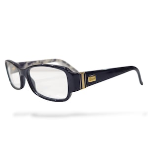 Könnte beinhalten: Schwarze rechteckige Brille mit klaren Gläsern. Die Fassungen haben einen goldenen Akzent und das Gucci-Logo. Die Bügel sind schwarz mit einem Muster in der Nähe der Scharniere. Die Brille ist vor einem weißen Hintergrund platziert.