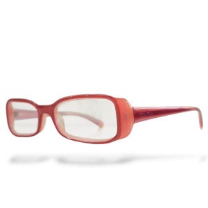 May include: Red rectangular eyeglasses with clear lenses. The frames are a solid red color with a slightly rounded shape. The temples are thin and extend to the ear pieces. The brand name is printed on the temple.