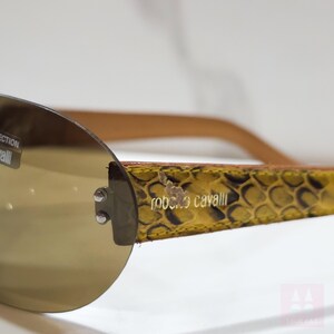 Peut inclure: Lunettes de soleil sans monture avec une teinte marron. La monture pr&eacute;sente un motif peau de serpent jaune et marron, avec le nom de la marque "roberto cavalli" visible. Les branches sont de couleur marron clair.