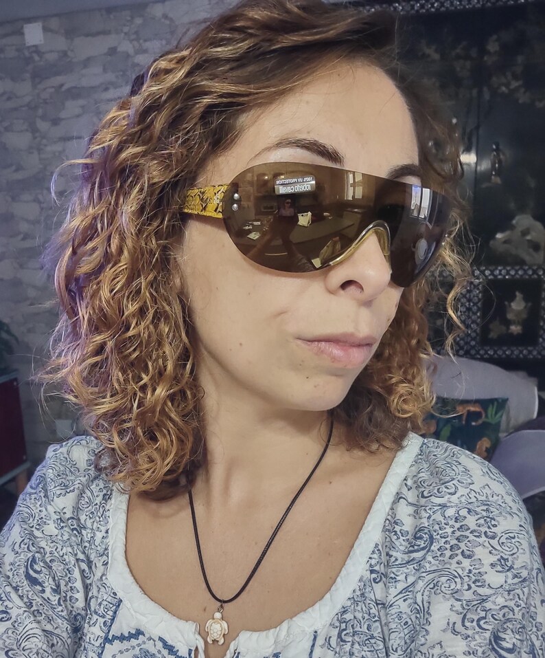 Peut inclure: Lunettes de soleil enveloppantes teint&eacute;es de marron. Les montures ont une bordure dor&eacute;e et un motif jaune et noir sur les branches. Les verres refl&egrave;tent l'environnement.