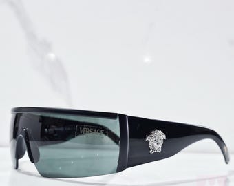 Puede incluir: Gafas de sol negras con un logotipo de Versace plateado y lentes de color gris oscuro. Las gafas tienen un diseño envolvente.