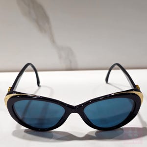 Puede incluir: Gafas de sol negras de ojo de gato con lentes azules. Las monturas son negras con detalles dorados en las patillas y un detalle dorado texturizado en la parte superior de la montura. Estilo retro y vintage.
