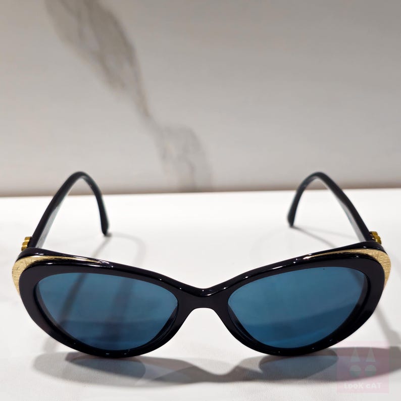 Puede incluir: Gafas de sol negras de ojo de gato con lentes azules. Las monturas son negras con detalles dorados en las patillas y un detalle dorado texturizado en la parte superior de la montura. Estilo retro y vintage.