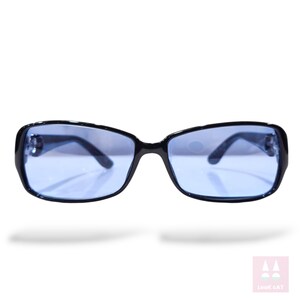 May include: Black-framed sunglasses with blue-tinted lenses. The rectangular frames have a slight cat-eye shape. The sunglasses are set against a white background.