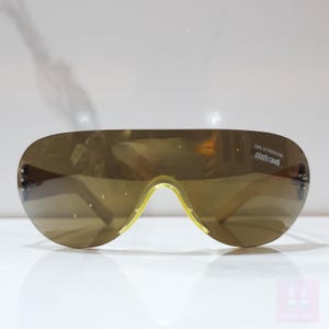 Peut inclure: Lunettes de soleil marron en forme d'&eacute;cran avec un pont et des branches jaunes. Les lunettes ont un design enveloppant incurv&eacute;. Le texte "100% UV Protection Roberto Cavalli" est imprim&eacute; sur la lentille.