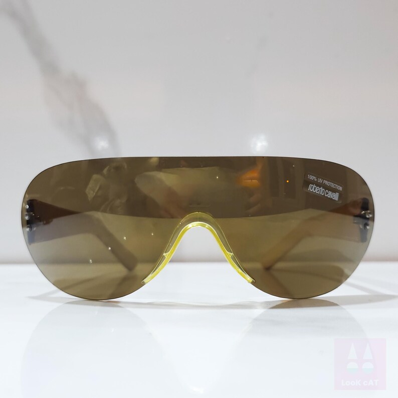 Peut inclure: Lunettes de soleil marron en forme d'&eacute;cran avec un pont et des branches jaunes. Les lunettes ont un design enveloppant incurv&eacute;. Le texte "100% UV Protection Roberto Cavalli" est imprim&eacute; sur la lentille.