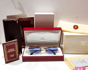 May include: A pair of gold-rimmed sunglasses with blue lenses in a red velvet box. The box has the words "must de Cartier" printed on it. The sunglasses are accompanied by a certificate of guarantee, a cleaning cloth, and a small pouch.