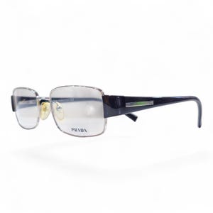 Peut inclure: Lunettes Prada à monture argentée et verres rectangulaires. Les branches sont noires avec un détail vert et argenté. Le logo Prada est imprimé sur la monture des verres.