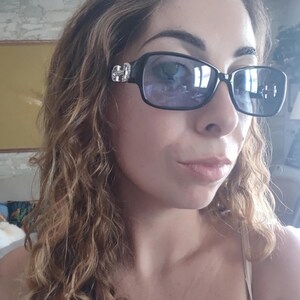 May include: Black rectangular sunglasses with silver embellishments on the temples. The lenses are a gradient purple color. The sunglasses are worn by a person with curly brown hair.
